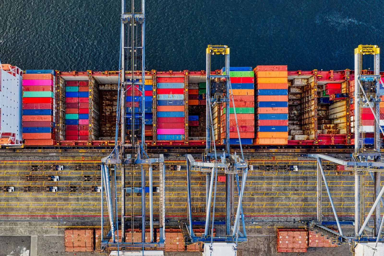 Containers op een vrachtschip naast havenkranen, recht van boven gezien.